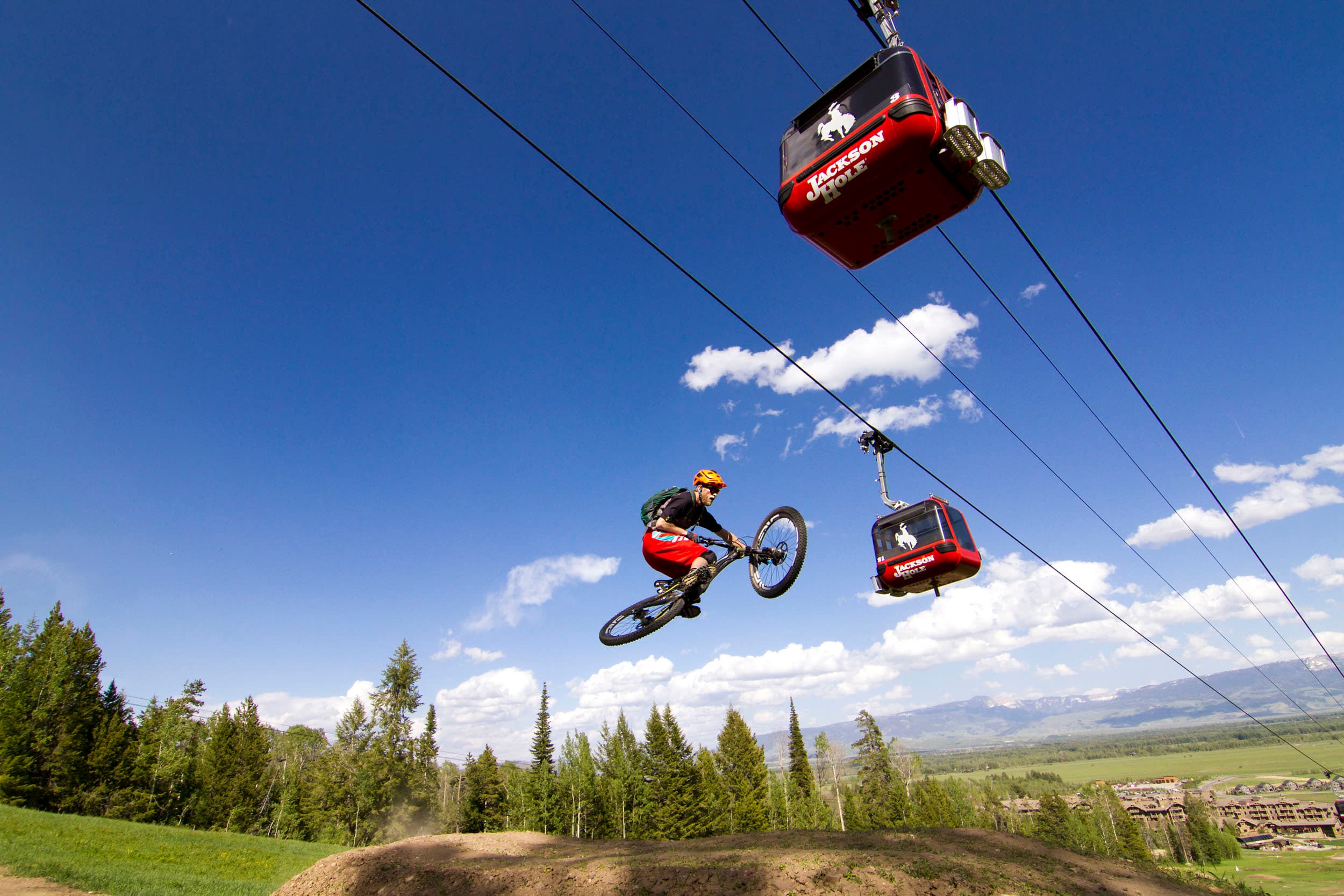 A person taking a jump at the mountain biking park with the Jackson Hole Mountain Resort tram passing above him.