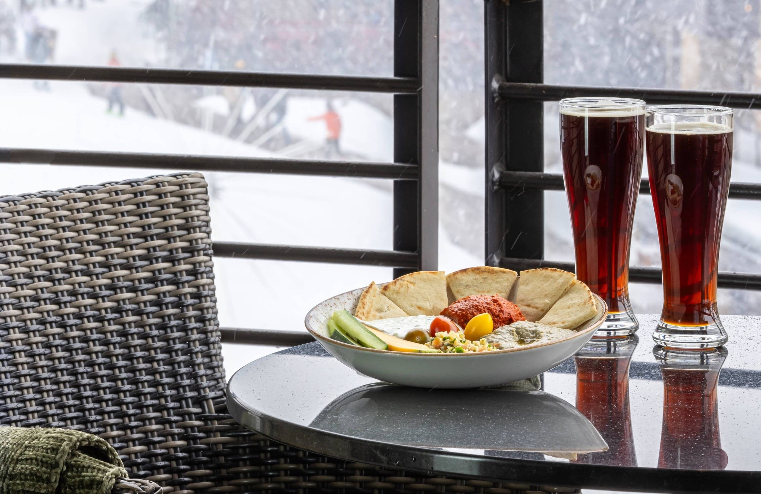 A plate of food in front of two beers on a table on the outdoor balcony at Hotel Terra Jackson Hole overlooking the snowy mountain.